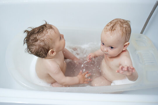 Twin babies splashing joyfully in a white plastic tub during their evening bath time as they engage in playful exploration together inside the home