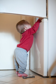 Toddler in a red shirt reaching up to open the white kitchen refrigerator door to find snacks in a bright home during the middle of the day time