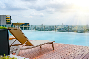 Fototapeta premium Serene rooftop infinity pool scene featuring a comfortable lounge chair on a wooden deck with a stunning panoramic view of the Bangkok cityscape at daytime
