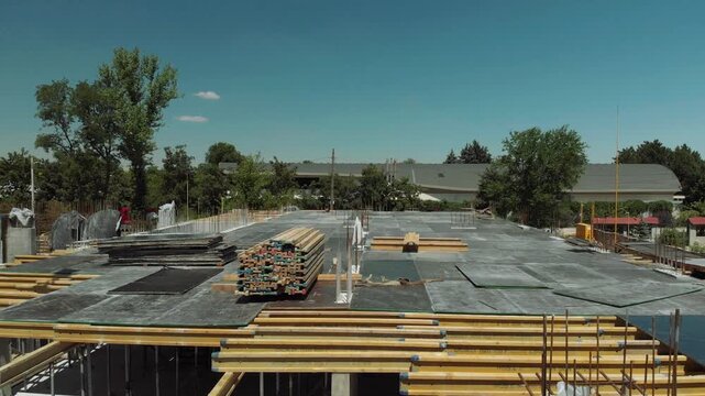 High-angle view shows organized storage of timber beams, steel reinforcement bars, concrete slabs, and formwork in outdoor yard with dust, shadows, and daylight