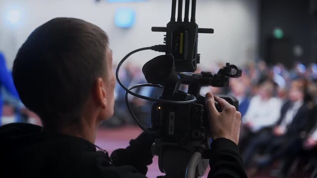 Professional cameraman filming a live conference with video camera. Event broadcasting, live streaming and video production with audience in the background at a business conference.