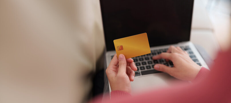 Person holding a credit card while shopping online on a laptop at home, representing digital payment, e-commerce purchase, cashless checkout, internet banking and convenient retail spending.