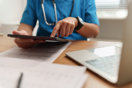 Female Doctor in Blue scrub using digital tabletand laptop while reviewing medical documents, representing clinical analysis, patient care, digital healthcare and modern medical technology.