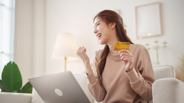 Happy Asian woman celebrating successful online purchase while holding credit card beside her laptop at home, representing digital payment, e-commerce checkout.