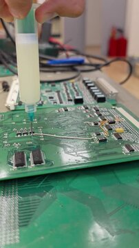 Syringe applying flux onto a green circuit board during electronics repair, close-up component replacement, precise soldering process.