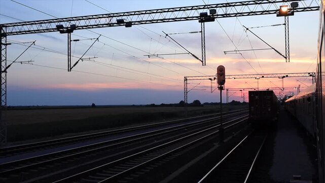 Railway tracks under a sunset sky. Electric lines stretch across the horizon. A train is parked on the side, surrounded by open fields.