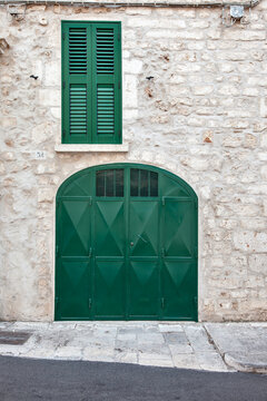A green metal double door with a diamond motif and matching shutters set into a rustic Lecce stone wall, Ostuni, Puglia, Italy.