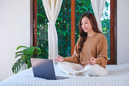 Portrait of a woman meditating or practicing yoga in lotus pose on bed in front of laptop computer