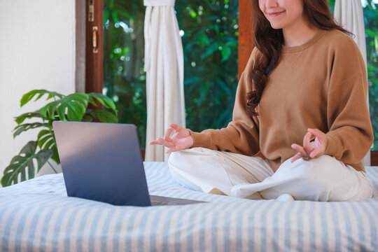 Closeup of a woman meditating or practicing yoga in lotus pose on bed in front of laptop computer