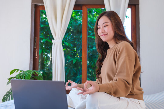 Portrait of a woman meditating or practicing yoga in lotus pose on bed in front of laptop computer