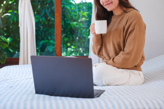 Closeup of a woman using and working on laptop computer while sitting and drinking coffee on the bed
