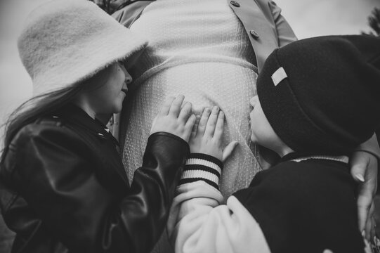 Cute children gently touching and kissing the belly of their pregnant mother outdoors during a family twin pregnancy photoshoot. Black and white photo.