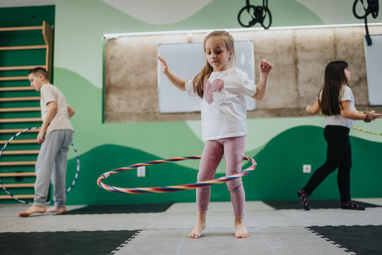 A young girl practices hula hooping barefoot in a school gym. Two other children in the background also play with hula hoops during a group activity.