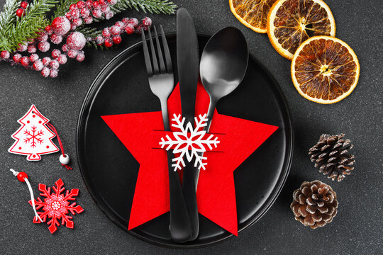 Holiday table setting with black cutlery arranged in red star shaped cutlery holder decorated with white snowflake, Christmas tree branch, ornaments and dried oranges on dark textured background