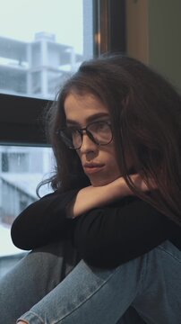 Burnout and emotional exhaustion, woman in glasses resting on folded arms by window. Loneliness, depression and mental health awareness. Vertical video.