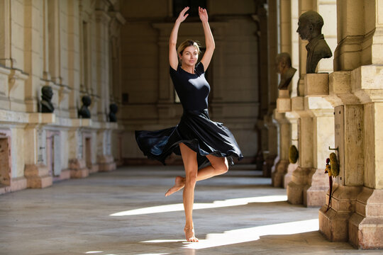 Dancer spinning in elegant black dress. Dancer barefoot inside historic interior. Full body dancer with raised arms. Graceful dancer in dynamic motion.