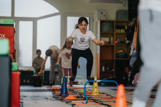 A young girl jumps over hurdles as part of a kids fitness obstacle course. Other children take turns in the indoor group activity at a school gym.
