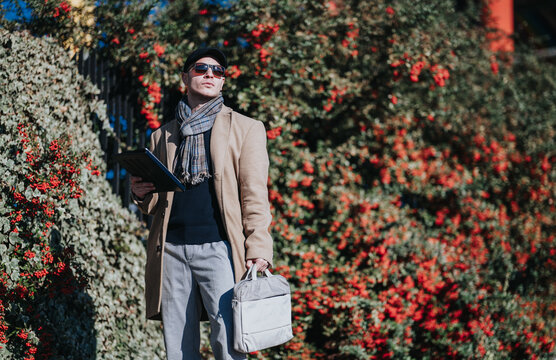A professional businessman wearing a coat and scarf holds a tablet and carries a laptop bag. He stands outdoors by red-berried foliage on a sunny autumn day.
