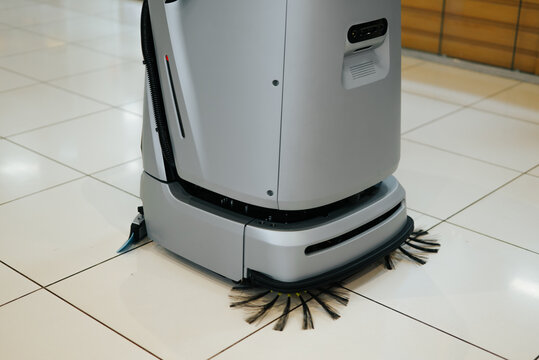 A cleaning robot with rotating brushes is positioned on a smooth tiled floor in a commercial space. The robot appears to be actively engaged in cleaning as its brushes rotate