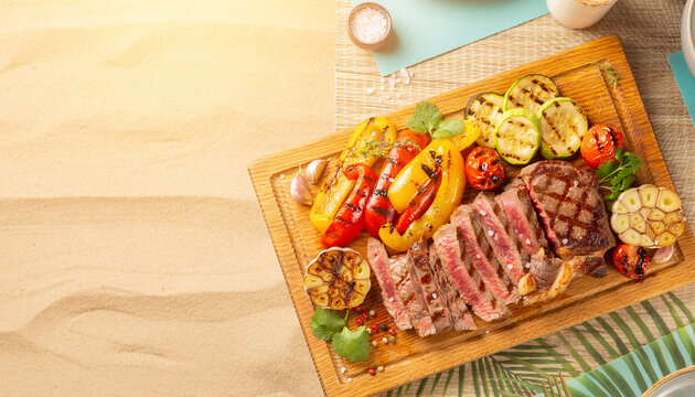 Grilled steak rests on wooden board. Bright peppers and zucchini add vibrant color. Garlic cloves and cherry tomatoes enhance the dish. Sliced steak shows perfect medium-rare doneness