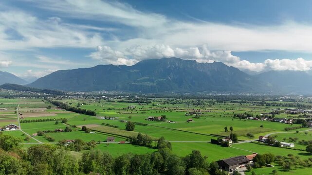 Aerial drone view of the flat Rhine valley floor with green fields and farms below a mountain massif in Switzerland