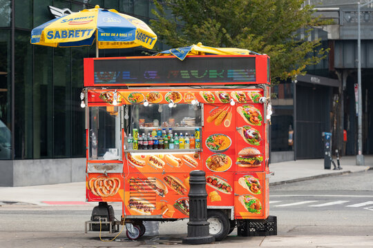 View of a traditional street food cart equipped with a colorful menu display and branded yellow umbrella located on an urban road in Manhattan, New York.