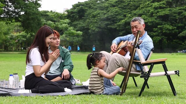 Asian kids painting in park. Asian family enjoy outdoor picnic with food and drinks in park sunny day, outdoor activitie with daughter father, mother grandfather. GrandFather playing acoustic guitar