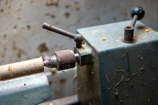 Old dusty wood lathe with a wooden workpiece mounted in the chuck, showing rusty metal parts, cobwebs and neglected woodworking machinery in a workshop.