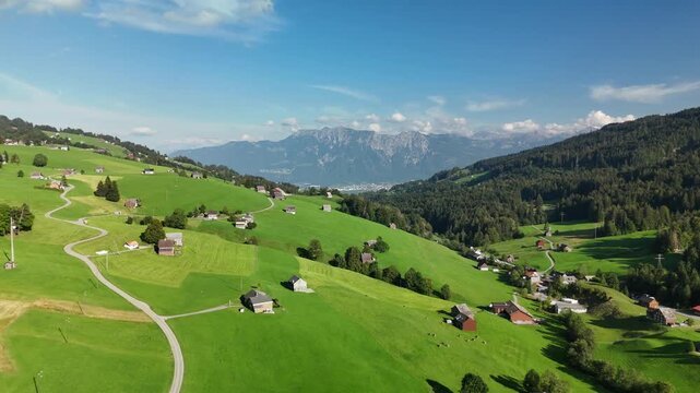 Aerial drone view of green alpine meadows with wooden barns and a village below rocky peaks in Switzerland
