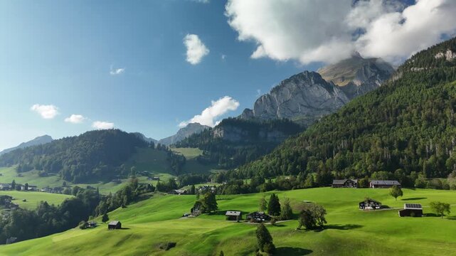 Aerial drone view of green alpine meadows with wooden barns and a village below rocky peaks in Switzerland