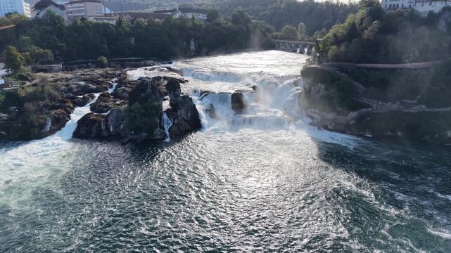 Wide aerial view of Rhine Falls in Switzerland, showing powerful cascades around massive rock formations, turbulent water below and a historic railway bridge in the background.