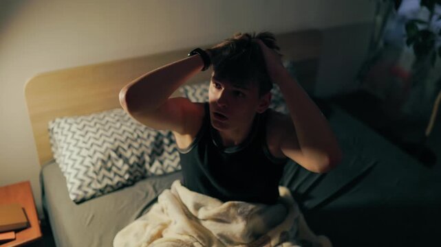 High angle of a stressed teenager rubbing his face in the dark bedroom.
