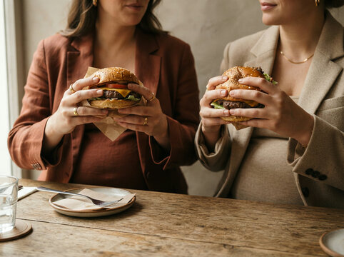 Two women in blazers holding paper wrapped burgers at a table in soft daylight