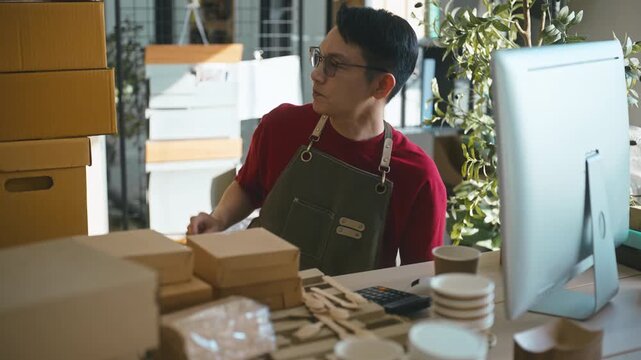 Asian male small business owner typing on a computer to check online customer orders, managing cardboard boxes and parcel delivery in a modern warehouse home office workspace.