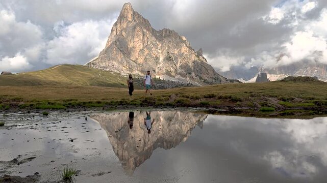 At Passo Giau in the Dolomites, Italy, two hikers stroll peacefully beside a serene pond, marveling at the breathtaking reflections of the majestic mountains. Natures beauty envelops them.