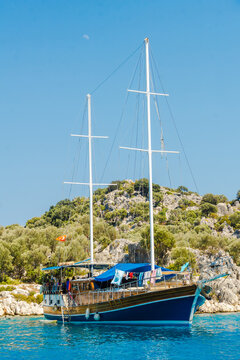 Two masted sailing yacht anchors near a rocky tree covered shoreline under a clear blue sky, Mediterranean sailing.