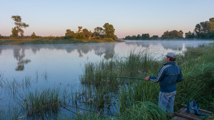 fisherman stands on the grassy riverbank holding a fishing rod during a calm, misty sunrise over the water © Александр Арендарь