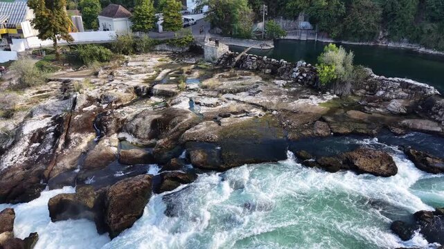 Close aerial view of Rhine Falls in Switzerland, showing powerful water channels rushing between massive exposed rocks with the historic railway bridge in the background.