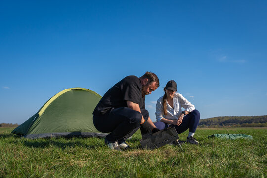 Couple crouching near a green tent, preparing a portable barbecue grill for cooking food while enjoying a sunny day of outdoor adventure and recreation in a field