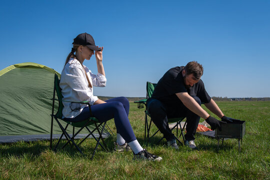 Man and woman squatting on a green grassy meadow, setting up a portable barbecue next to a small green tent under a clear blue sky, enjoying an outdoor adventure