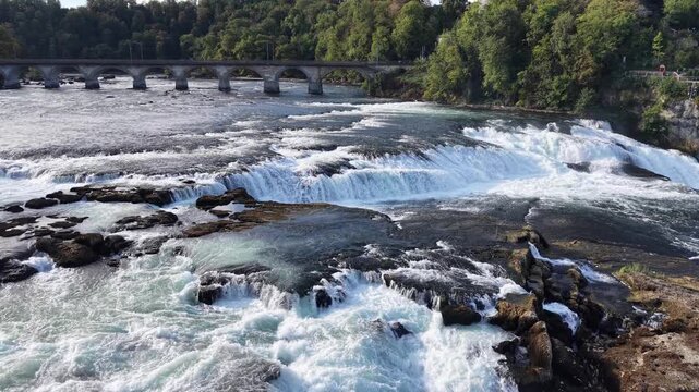 Close aerial view of Rhine Falls in Switzerland, showing powerful water channels rushing between massive exposed rocks with the historic railway bridge in the background.