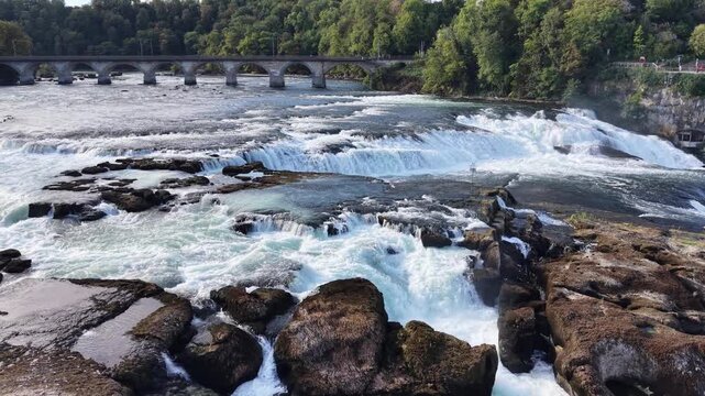 Close aerial view of Rhine Falls in Switzerland, showing powerful water channels rushing between massive exposed rocks with the historic railway bridge in the background.