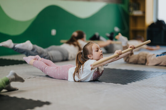A young girl and other kids lie on mats reaching forward with wooden bars. Children participate in a group floor exercise class focused on balance and strength.
