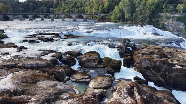 Close aerial view of Rhine Falls in Switzerland, showing powerful water channels rushing between massive exposed rocks with the historic railway bridge in the background.