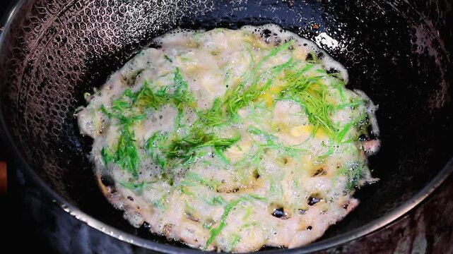 Close up of a Thai cha om omelet frying in bubbling oil inside a dark wok in Thailand, showing climbing wattle leaves and egg during a street food cooking process at night.