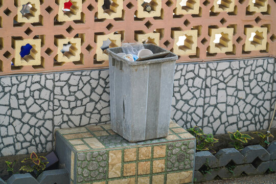 Outdoor trash bin filled with plastic waste against a decorative mosaic wall with geometric patterns and small potted plants nearby, elegantly showcasing background.