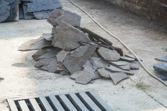 Stack of rough cut stone slabs piled on concrete ground near drainage grate and brick wall, suggesting construction or demolition site with exposed aggregate material and outdoor work environment.