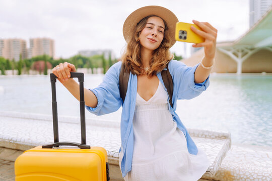 Young female tourist wearing hat records vlog on phone while sitting on bench with yellow suitcase. Woman is relaxing and taking selfie outdoors. Concepts: tourism, blogging, and urban life.