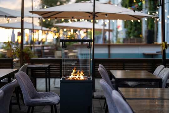 Cozy open-air restaurant terrace with sofas, umbrellas and modern gas fireplace inside glass box, glass patio heater with burning flame beside comfortable dining tables at outdoor cafe settings.