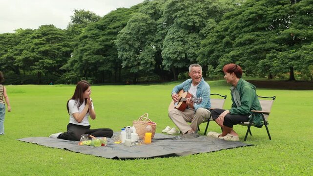 Asian kids painting in park. Asian family enjoy outdoor picnic with food and drinks in park sunny day, outdoor activitie with daughter father, mother grandfather. GrandFather playing acoustic guitar